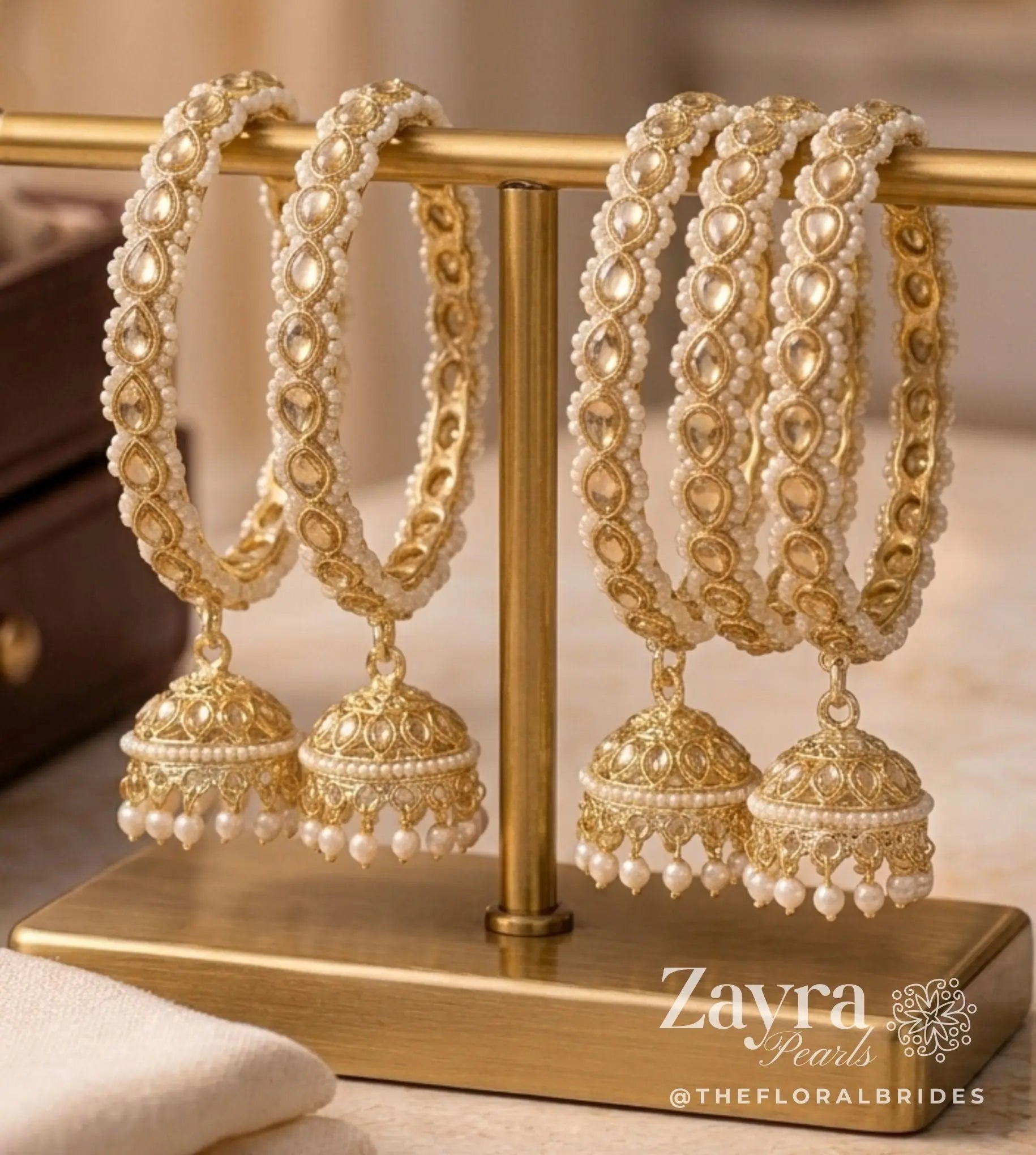 Zayra Pearls royal gold Kundan kangan bangles in a pair featuring hanging jhumkas bordered with tiny white seed pearls on a gold T-bar display