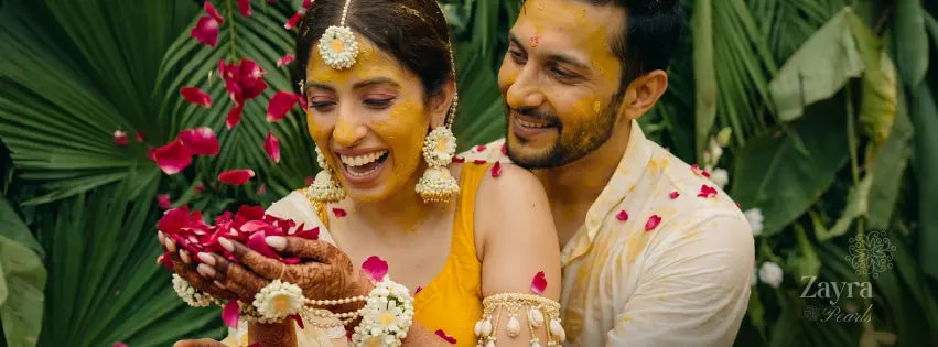 A smiling bride and groom surrounded by lush green tropical leaves, wearing haldi jewellery set by zayra pearls.