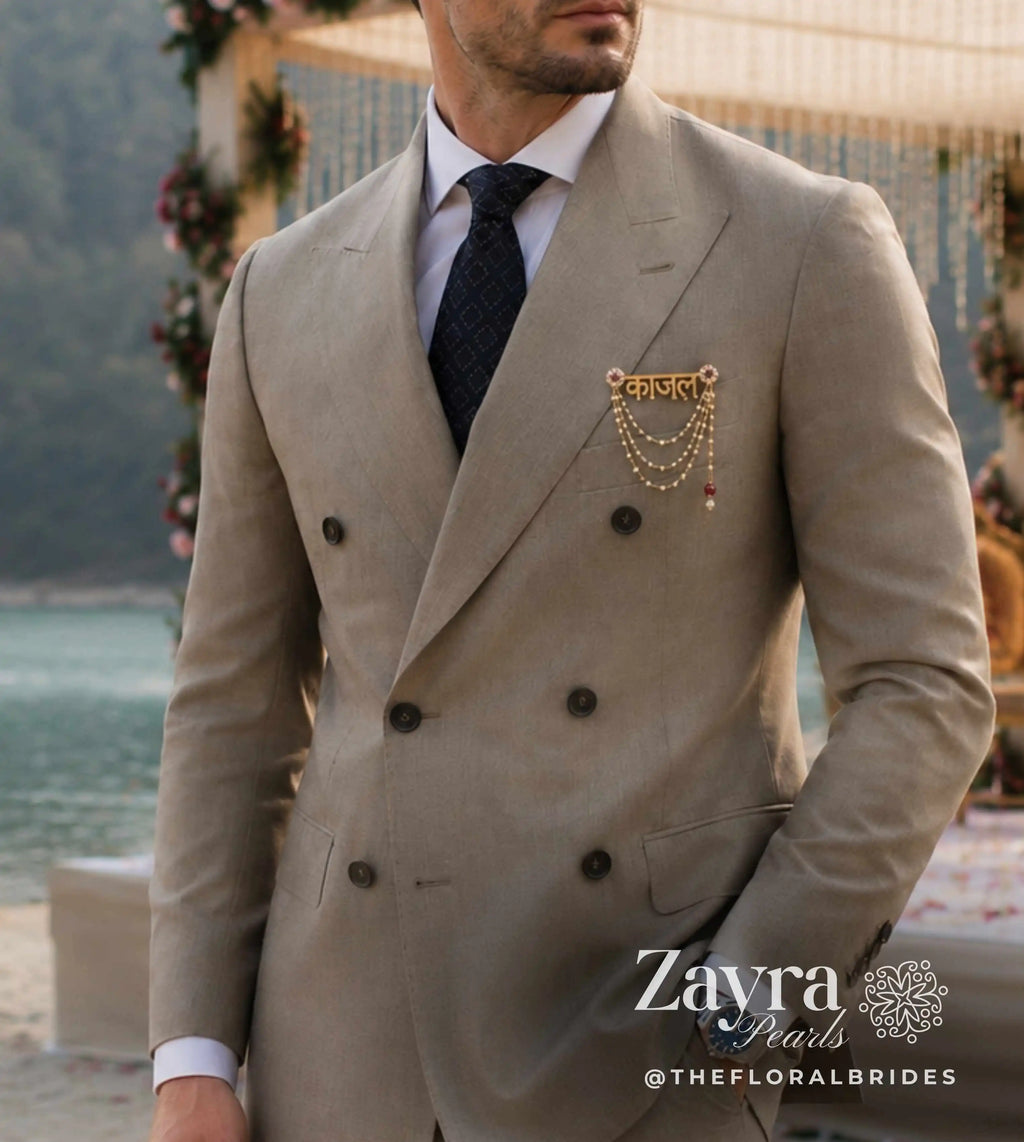 A man wearing a light beige double-breasted suit jacket with a gold chain brooch from zayra pearls on the lapel, paired with a white shirt and dark tie, standing outdoors near a beach and floral arch.