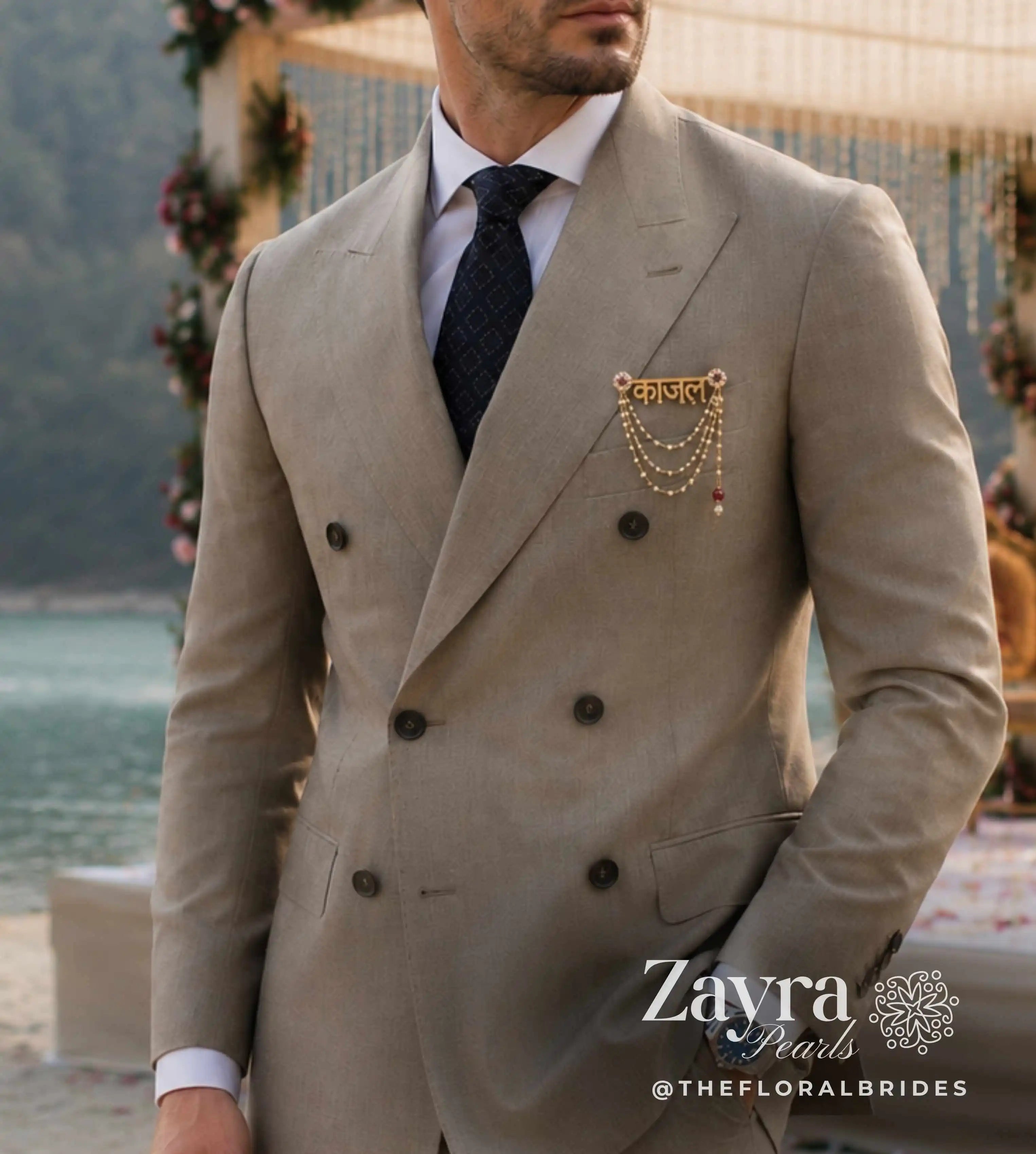 A man wearing a light beige double-breasted suit jacket with a gold chain brooch from zayra pearls on the lapel, paired with a white shirt and dark tie, standing outdoors near a beach and floral arch.