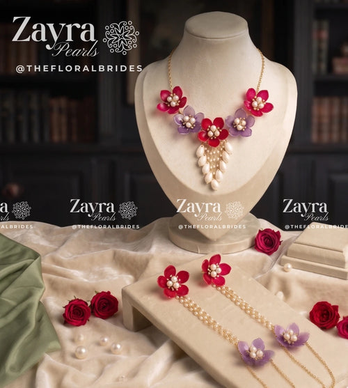 A jewelry set displayed on a cream mannequin bust and trays, featuring red and purple flower-shaped pearl necklaces, matching earrings, and pearl strands with gold-tone chains. The set is styled with red roses and draped cream fabric against a dark, blurred background.