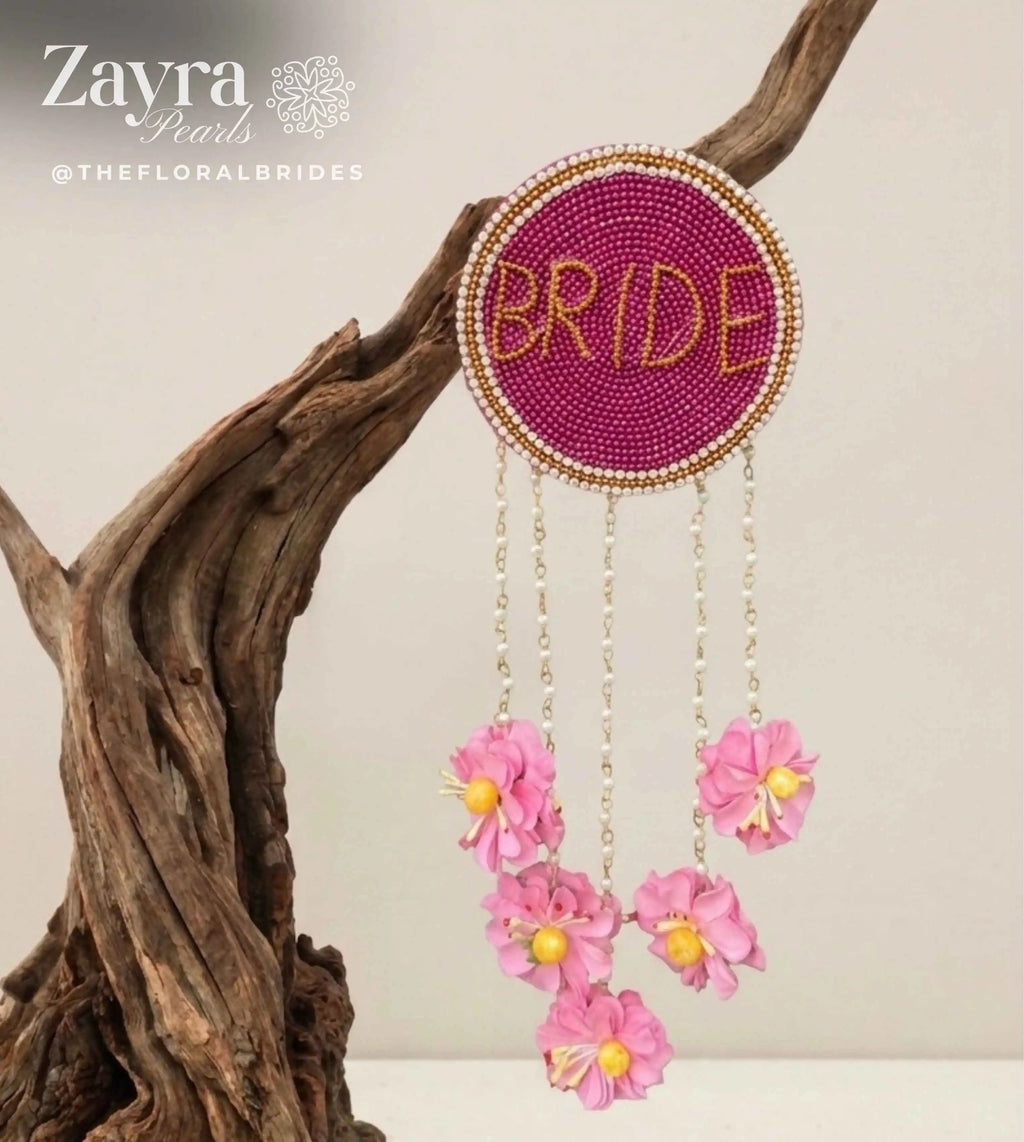 Personalized Bridal Hair Accessory | Zayra Pearls - A circular beaded wall hanging with the word 'BRIDE' in gold beads on a magenta background, suspended from a piece of driftwood. Three dangling chains hang pink flowers with yellow centers.