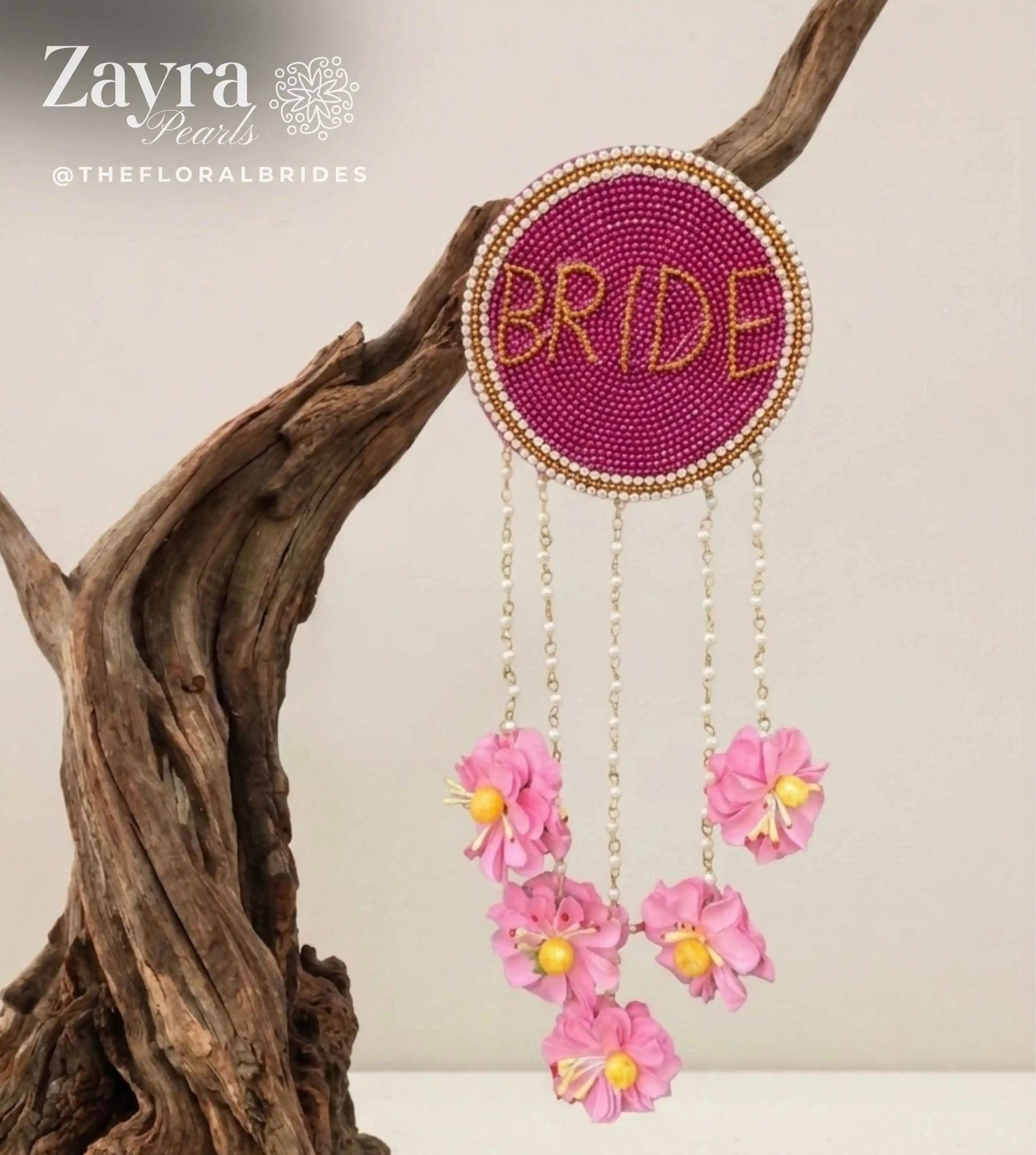 Personalized Bridal Hair Accessory | Zayra Pearls - A circular beaded wall hanging with the word 'BRIDE' in gold beads on a magenta background, suspended from a piece of driftwood. Three dangling chains hang pink flowers with yellow centers.