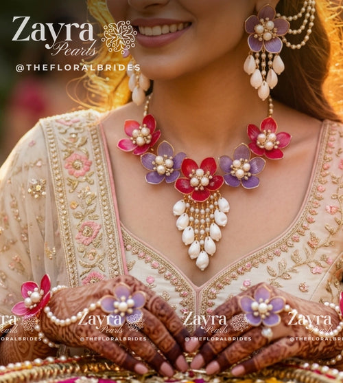 A close-up lifestyle shot of a woman wearing a Zayra Pearls bridal jewelry set, featuring a floral necklace with red, purple, and white flowers, matching drop earrings, and floral henna designs on her hands, set against a warm, sunlit background.
