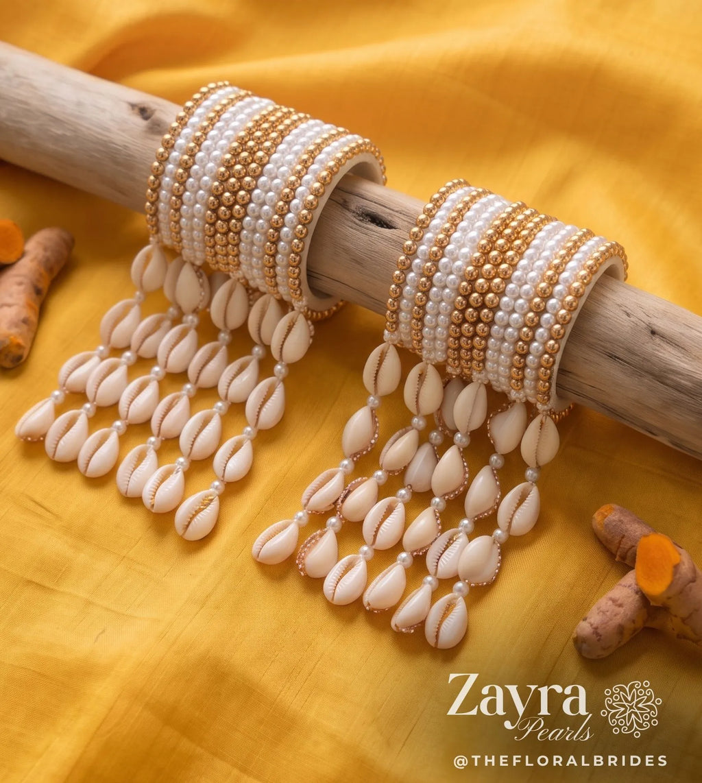 ZAYRA PEARLS beaded bangles with dangling cowrie shell charms, displayed on a rustic wooden branch over a bright yellow fabric backdrop. Turmeric roots are scattered around as decorative props.