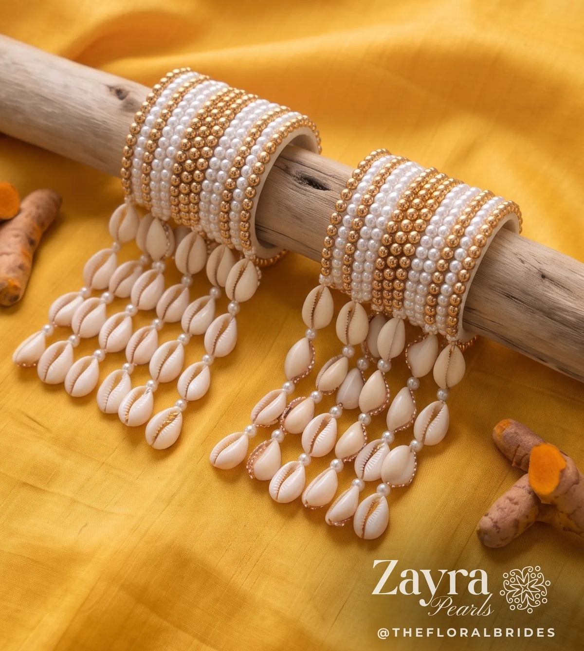 ZAYRA PEARLS beaded bangles with dangling cowrie shell charms, displayed on a rustic wooden branch over a bright yellow fabric backdrop. Turmeric roots are scattered around as decorative props.