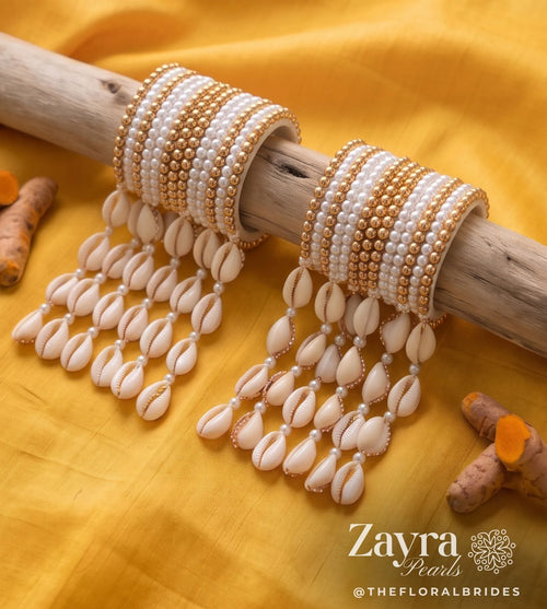 ZAYRA PEARLS beaded bangles with dangling cowrie shell charms, displayed on a rustic wooden branch over a bright yellow fabric backdrop. Turmeric roots are scattered around as decorative props.