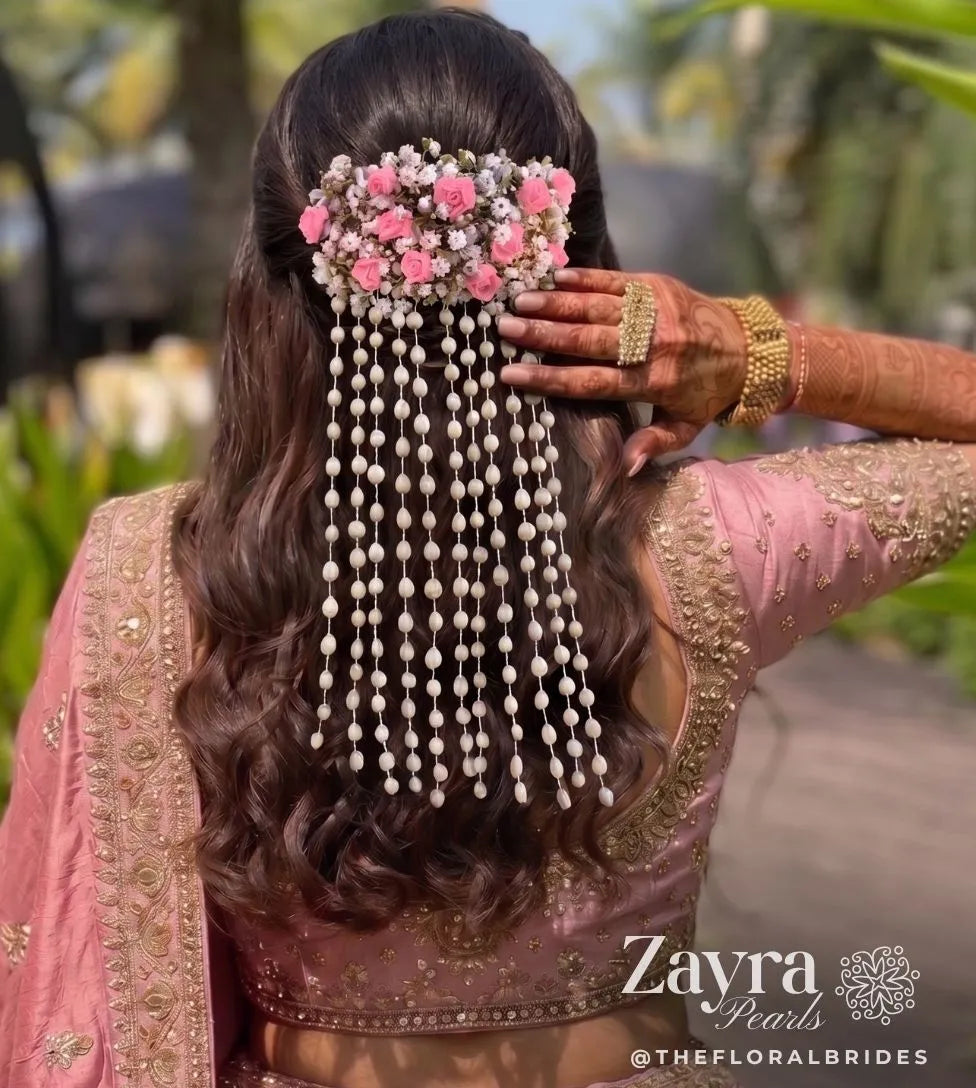 Zayra pearls model shown from behind wearing a pink floral hair accessory with long dangling pearl strands, styled with a pink embroidered blouse and jewelry in an outdoor setting. The brand name 'Zayra Pearl' is visible as a watermark at the bottom right.