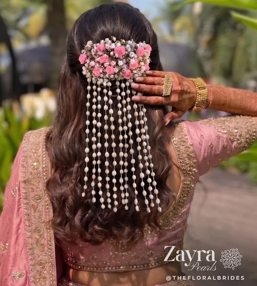 Zayra pearls model shown from behind wearing a pink floral hair accessory with long dangling pearl strands, styled with a pink embroidered blouse and jewelry in an outdoor setting. The brand name 'Zayra Pearl' is visible as a watermark at the bottom right.