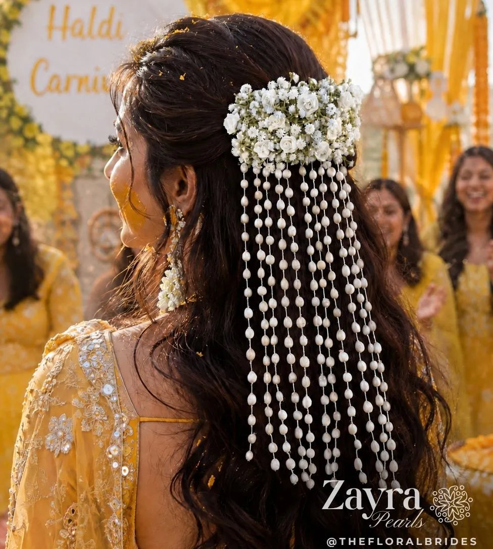 A woman in a yellow traditional outfit shown from the back, wearing a decorative floral hairpiece from Zayra pearls with long dangling pearl-like strands. Bride wearing floral bridal juda accessories with cascading white pearl strands for Haldi ceremony