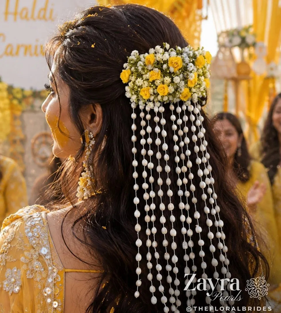 zayra pearls model ln in a yellow traditional outfit shown from the back, wearing a floral hair accessory with long dangling pearl bead strands. The hair accessory features yellow and white flowers at the top and cascading white bead fringes below. 