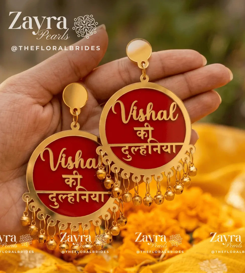 A pair of gold-plated round drop earrings with red enamel inlays and Hindi text reading 'Vishal ki Dulihaaniya,' held in a person's hand against a blurred outdoor background with yellow flowers. Brand watermarks for Zayra Pearls are visible.