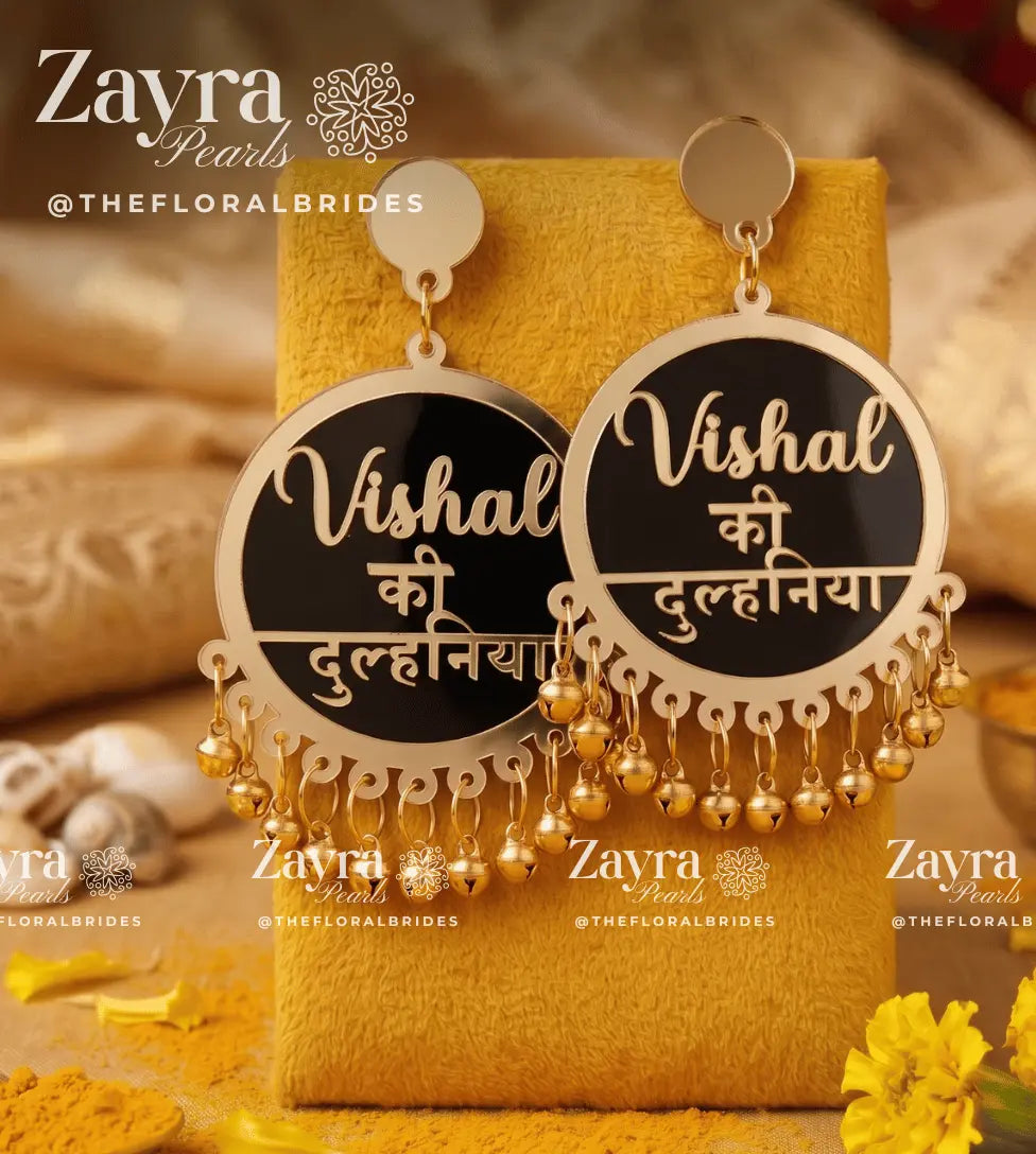 A pair of gold-toned statement earrings displayed on a yellow fabric stand. Each earring has a round black face with gold script text reading 'Vishal ki Dulhiniya' and dangling gold bell chains.