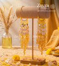 Yellow and gold beaded dangle earrings displayed on a wooden stand, with decorative dried grass in a glass vase and yellow flowers in the background. Watermarks for 'Zayra Pearls @THEFLORALBRIDES' are visible.