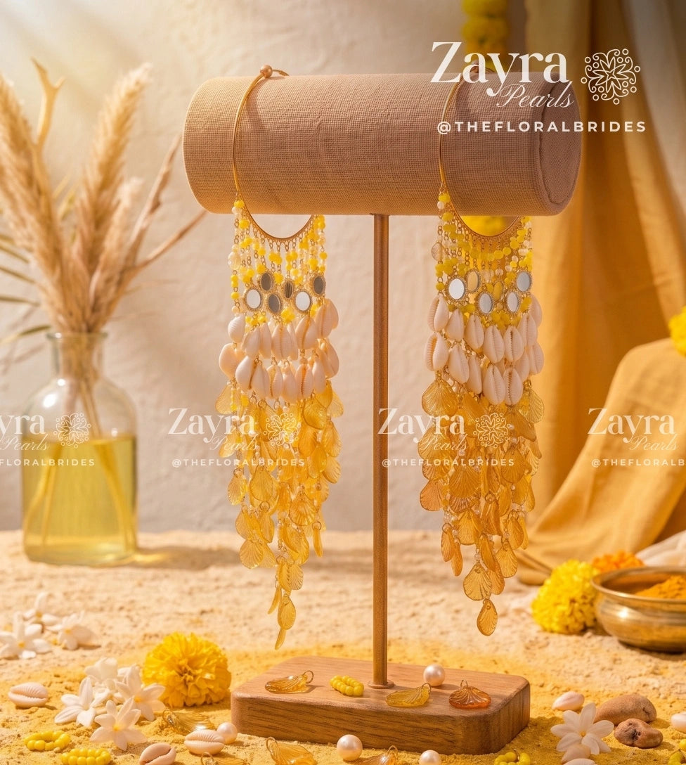 Yellow and gold beaded dangle earrings displayed on a wooden stand, with decorative dried grass in a glass vase and yellow flowers in the background. Watermarks for 'Zayra Pearls @THEFLORALBRIDES' are visible.