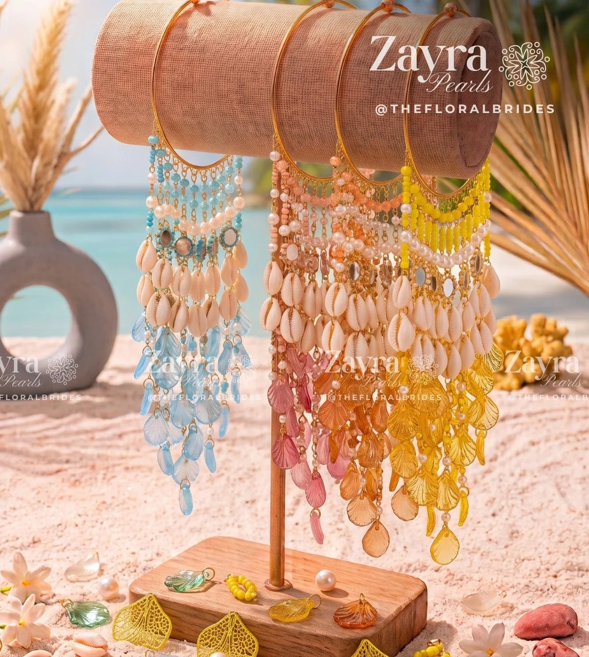 Three long beaded and shell fringe earrings displayed on a wooden stand in a beach-themed setting with sand, palm leaves, and turquoise water in the background. The earrings feature pastel blue, peach, pink, and yellow tones with shell and bead cascades.
