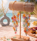 Colorful dangling earrings displayed on a wooden stand in a beach-themed setting with sand Handcrafted haldi carnival bangels with multicolor yellow blue pink and orange resin petals and cowrie by zayra pearls