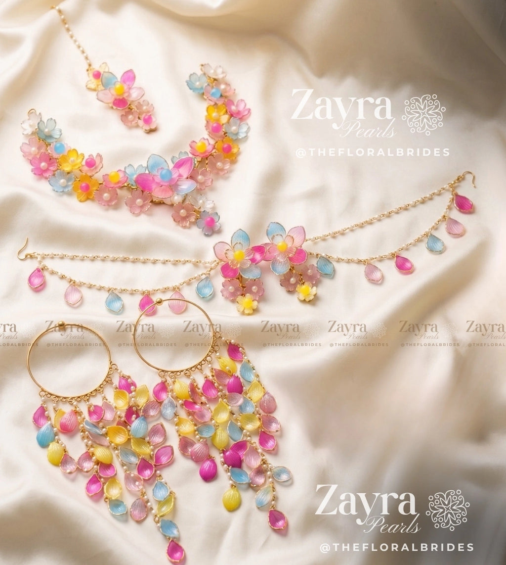 A jewelry set from Zayra Pearls featuring a gold chain necklace, matching choker, earrings, and a maang tikka, all decorated with colorful pastel flower and teardrop beads in pink, yellow, and blue, displayed on a cream satin fabric background.