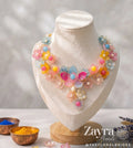 A colorful floral statement necklace displayed on a cream-colored fabric jewelry bust. The necklace features multiple layers of translucent flower petals in pastel shades of pink, yellow, blue, and orange, accented with small pearl-like beads.