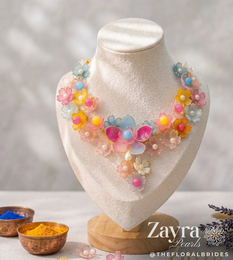 A colorful floral statement necklace displayed on a cream-colored fabric jewelry bust. The necklace features multiple layers of translucent flower petals in pastel shades of pink, yellow, blue, and orange, accented with small pearl-like beads.