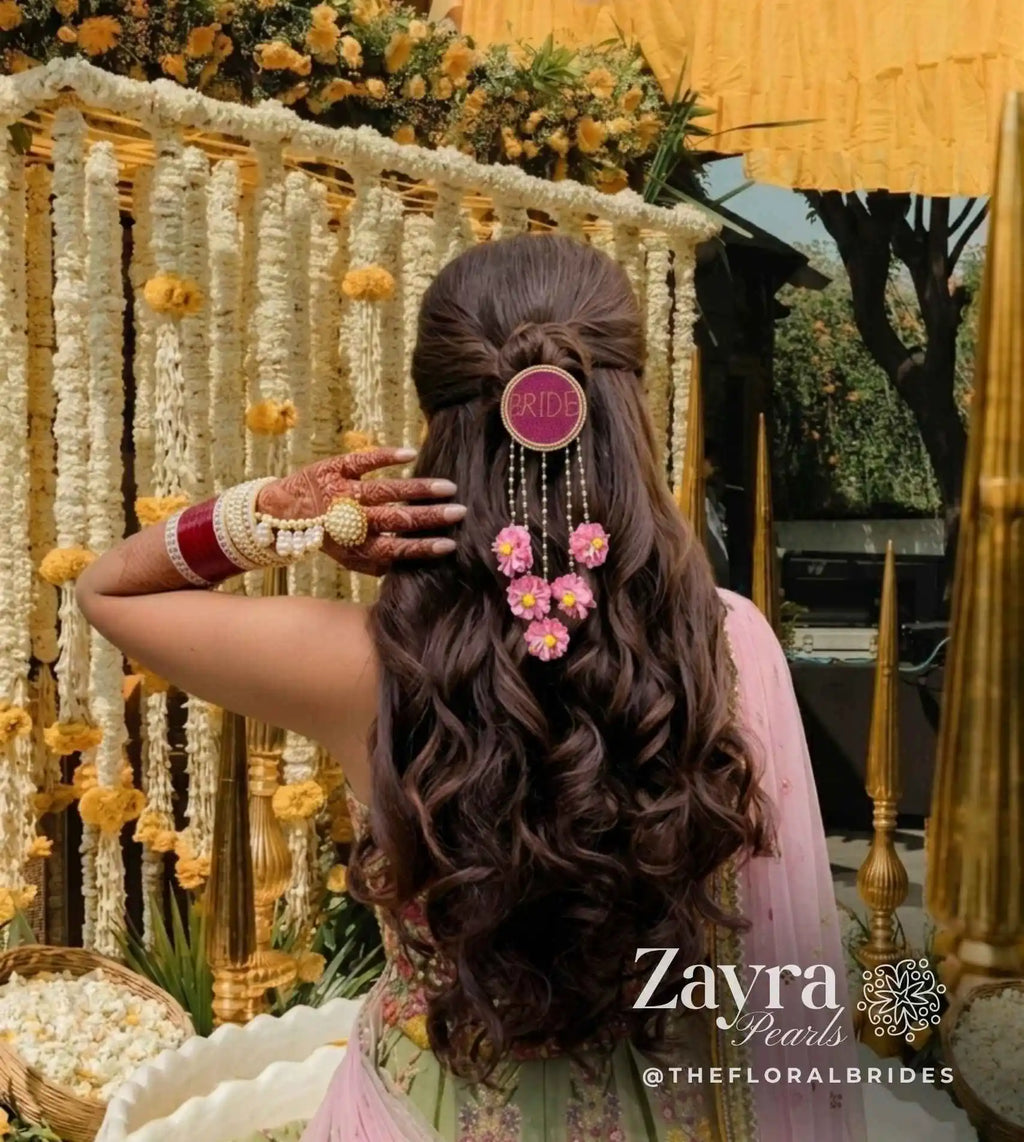 pink -Personalized Bridal Hair Accessory | Zayra Pearls woman shown from behind wearing a long, dark brown wavy hairstyle with a half-up bun, decorated with a pink circular hair pin and a floral hair clip. She is wearing a pink and green traditional outfit and has red and white bangles on her hand.