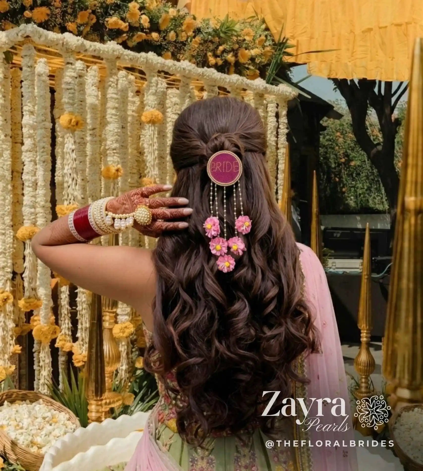 pink -Personalized Bridal Hair Accessory | Zayra Pearls woman shown from behind wearing a long, dark brown wavy hairstyle with a half-up bun, decorated with a pink circular hair pin and a floral hair clip. She is wearing a pink and green traditional outfit and has red and white bangles on her hand.