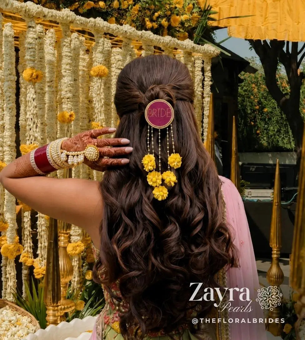 yellow Personalized Bridal Hair Accessory | Zayra Pearls woman viewed from behind wearing a decorative hair accessory with long dark brown curled hair, a pink circular hair pin reading BRIDE, and dangling yellow flower charms. She is posed in front of a floral garland backdrop with gold poles and outdoor greenery visible.