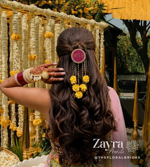 yellow Personalized Bridal Hair Accessory | Zayra Pearls woman viewed from behind wearing a decorative hair accessory with long dark brown curled hair, a pink circular hair pin reading BRIDE, and dangling yellow flower charms. She is posed in front of a floral garland backdrop with gold poles and outdoor greenery visible.