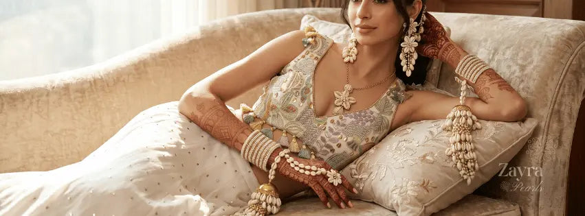 model reclining on a beige sofa wearing zayra pearls haldi shell jewelry, including a heavily embellished cream and gold embroidered dress, matching jewelry pieces, and stacked bangles.